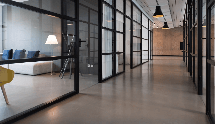 A hallway in an office building, looking down the corridor with two offices on each side. The offices have open doorways with glass walls, and through the glass, one can see a wooden table, a lamp, and a white couch with blue pillows inside one of the offices on the left.