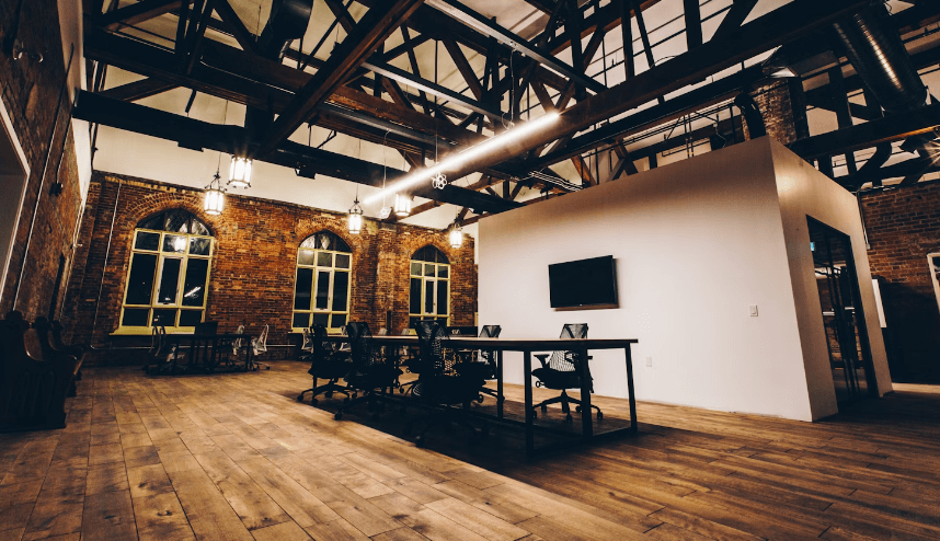 A warm office space with brick walls, wooden floors, and a high open ceiling with light fixtures. The room is spacious and clean, featuring a wooden table in the center surrounded by office chairs.